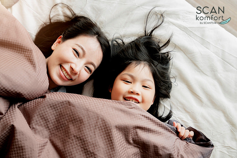 Two people lying in bed under brown blanket, smiling and relaxed in softly lit cozy bedroom.